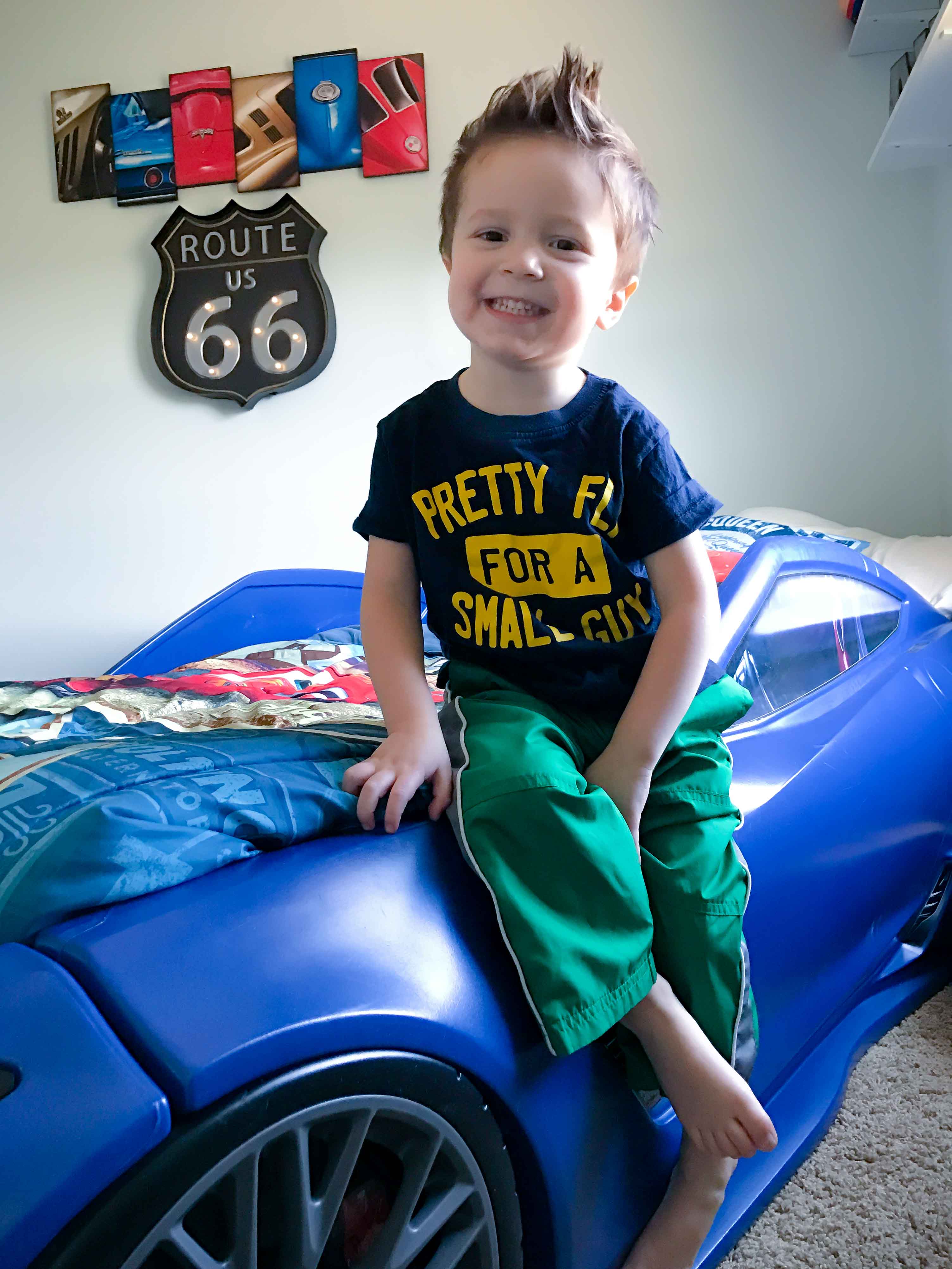A toddler boy sitting on a blue race car bed. 